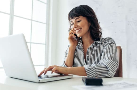 An image of a person on the phone while sitting in front of a laptop and smiling.